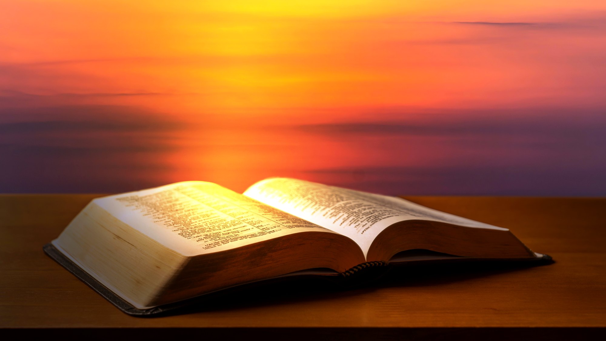 An open Bible resting on a wooden table against a sunset sky