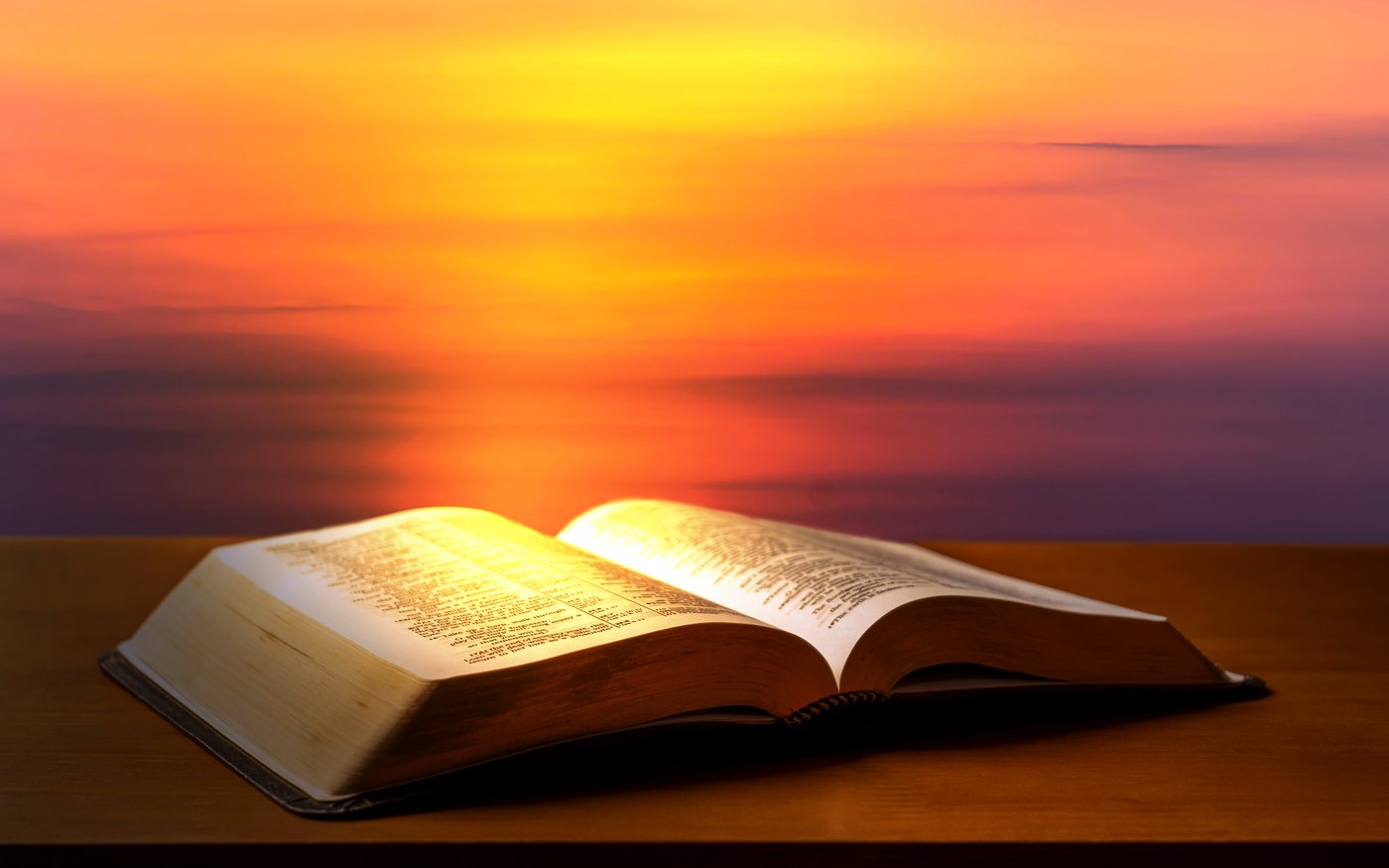 An open Bible resting on a wooden table against a sunset sky